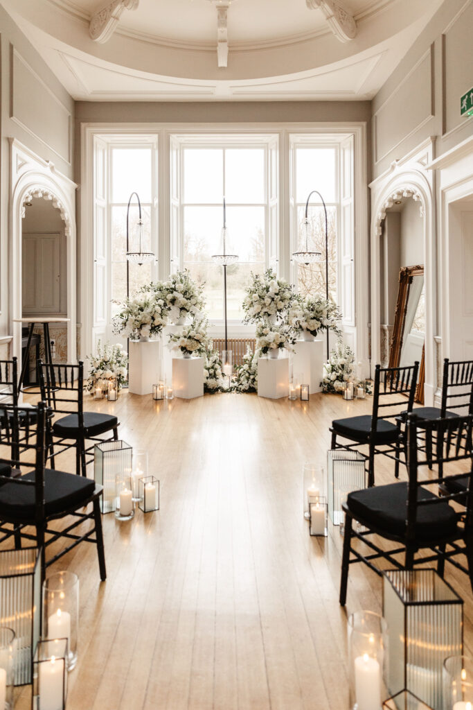 Modern indoor wedding ceremony setup with white floral pedestals, black chairs, and candles at Netherbyres House.