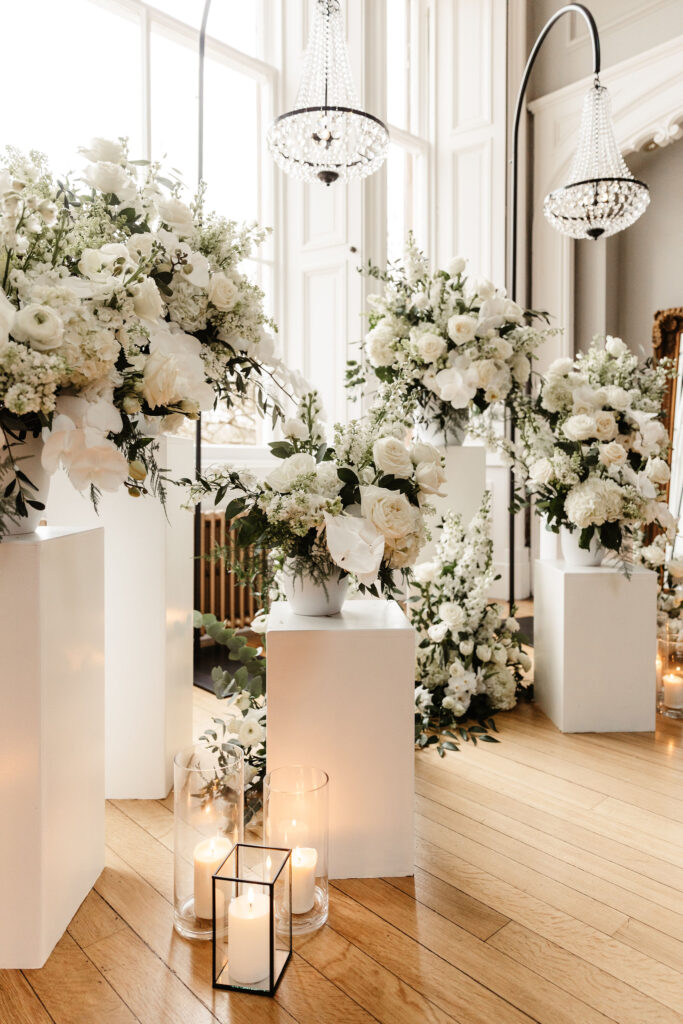 White floral arrangements on modern pedestals with candles and chandeliers in a light-filled room at Netherbyres House.