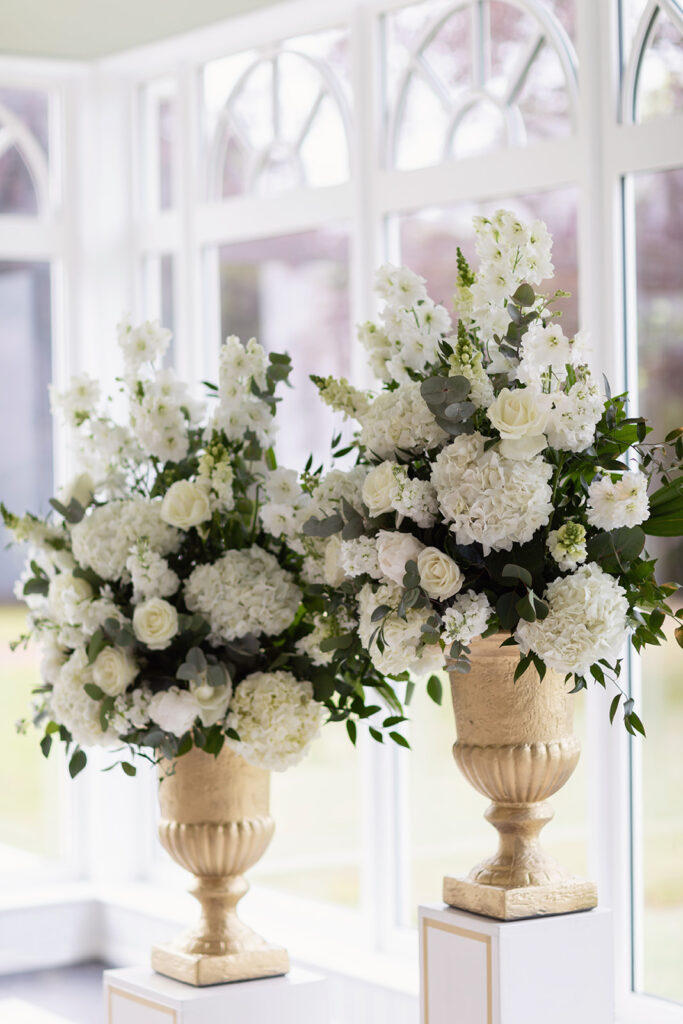 Two large floral arrangements in stone urns with white hydrangeas, roses, and delphiniums by bright windows.